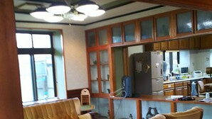 A spacious living room with a wooden floor, a sofa, and a coffee table. There is a kitchen area with wooden cabinets and a refrigerator. - Kawaraya-Ikinpata (Motobu)