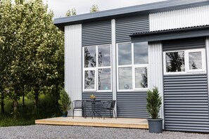 Un bâtiment moderne doté de grandes fenêtres, d’un petit espace extérieur aménagé avec des chaises et une table, et d’une plante en pot.