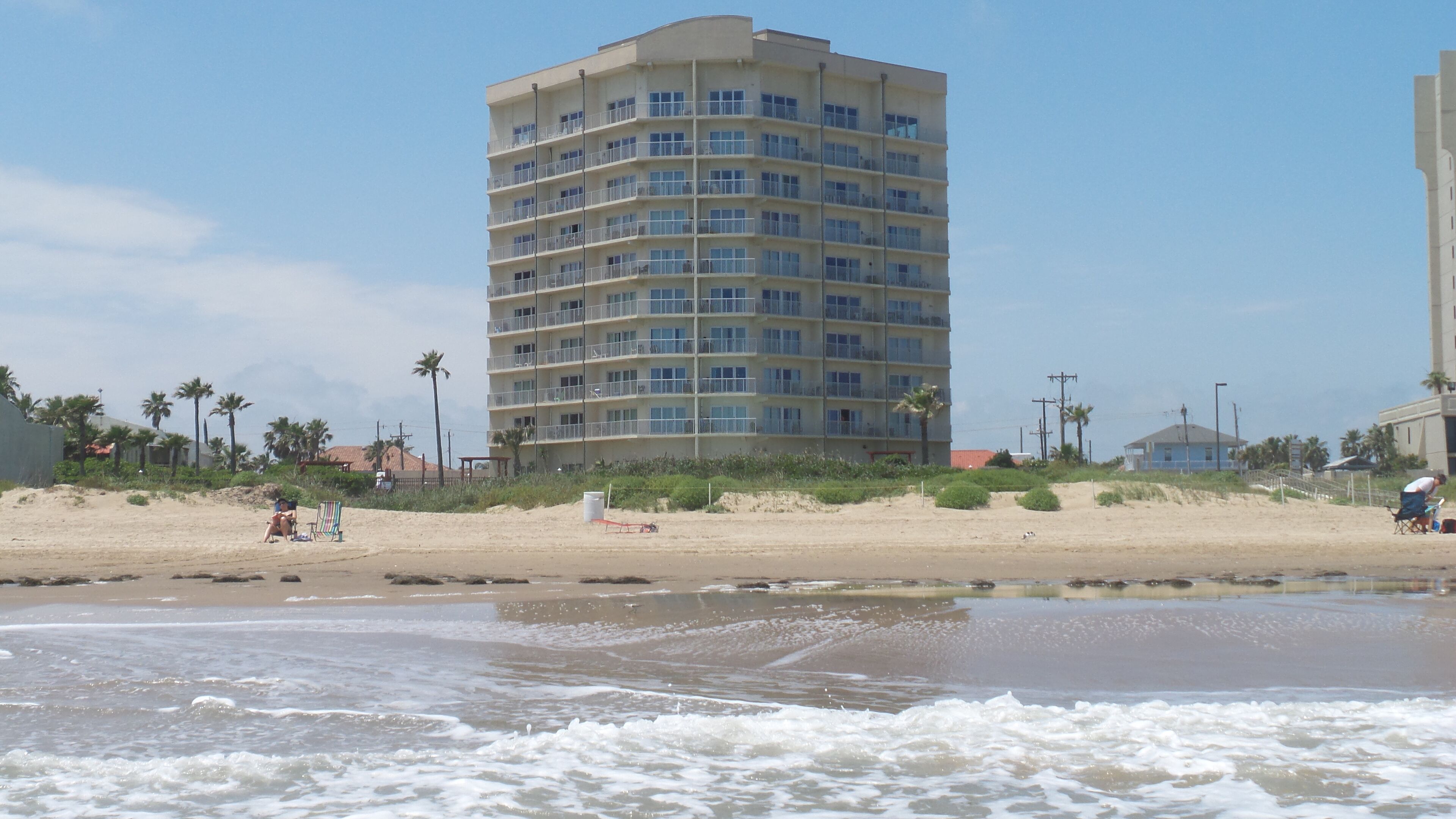 A multi-story hotel with balconies, located near a beach with palm trees and a sandy shoreline.