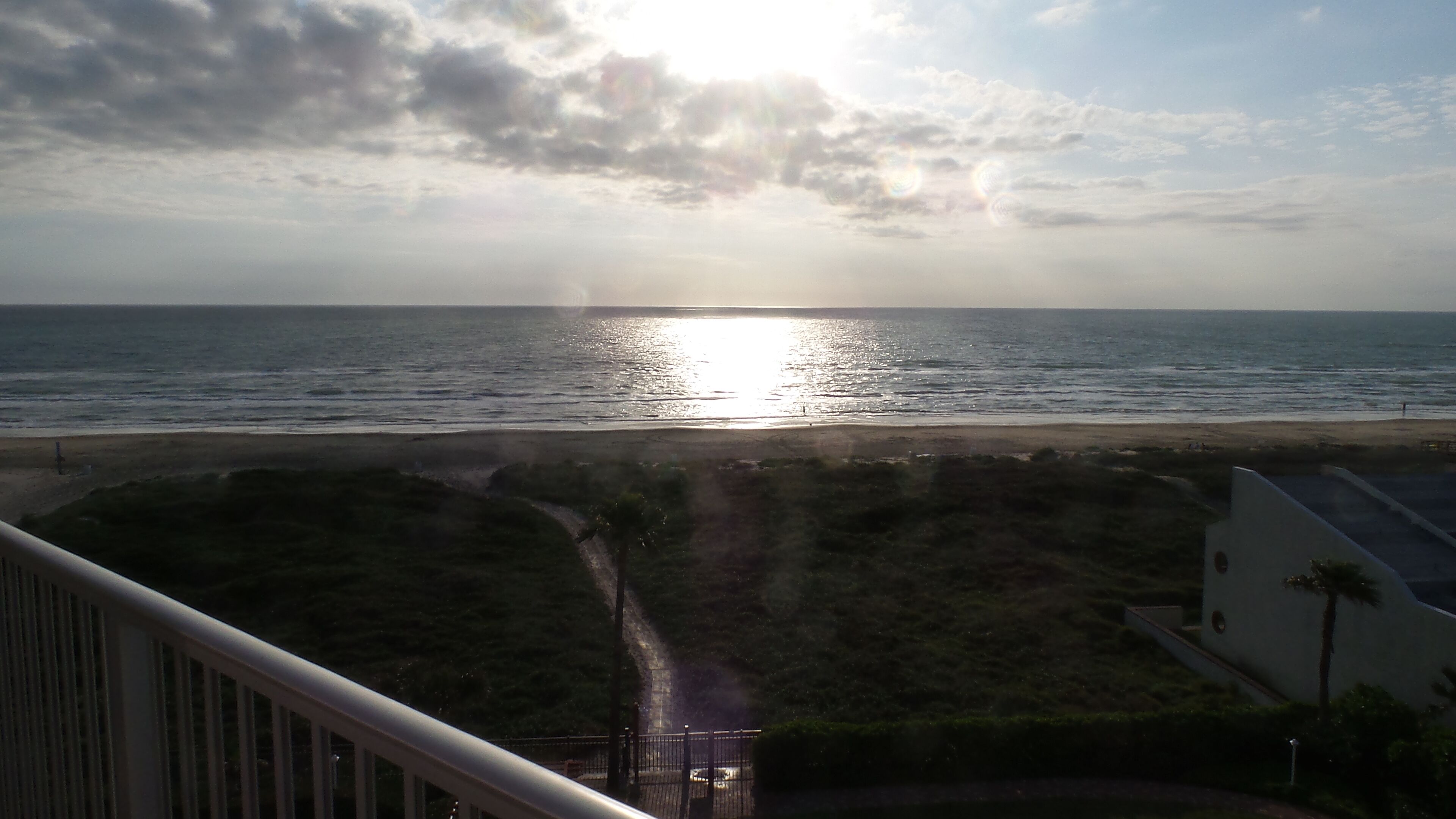 A view of the ocean with the sun reflecting on the water, a beach in the foreground, and a building with a fenced area.
