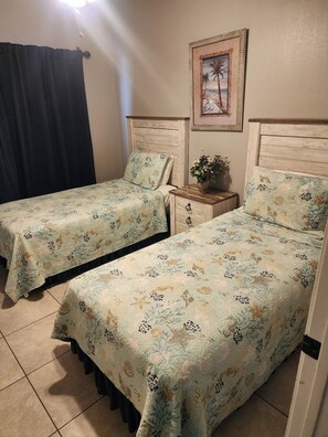 Una habitación con dos camas, cada una con ropa de cama estampada con motivos florales, una mesita de noche con una planta y un cuadro en la pared.