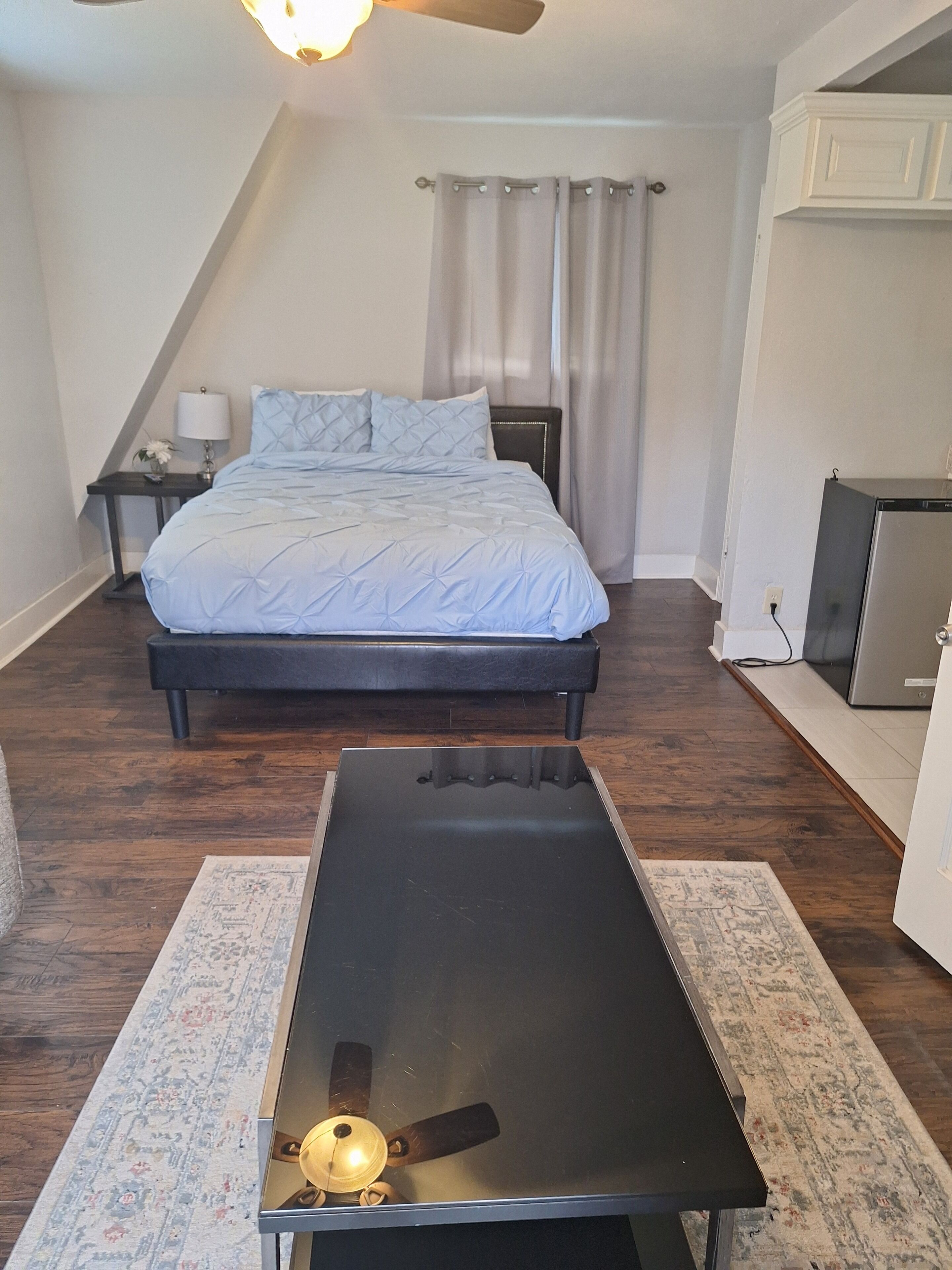 Une chambre à coucher comprenant un lit, un bureau, un ventilateur de plafond, une table de chevet, une lampe et un réfrigérateur.