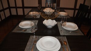 Mesa de comedor dispuesta con platos blancos, cubiertos y copas de vino, rodeada de sillas.