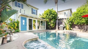 A swimming pool with a hot tub, surrounded by a patio with chairs and tables, and a house with a blue door and windows. - Naples Island Pool House (Long Beach)