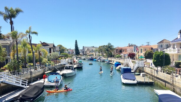 A marina with boats docked along the water, a pedestrian bridge, and houses in the background. - Naples Island Pool House (Long Beach)