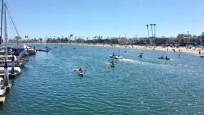 A marina with boats docked, a beach with people, and buildings in the background. - Naples Island Pool House (Long Beach)