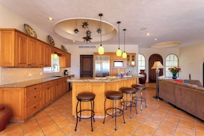 A kitchen with wooden cabinets, a central island with stools, pendant lighting, a refrigerator, and a sofa in the living area. - Villa Agave Azul | 5BR Ocean View Villa | Pool, Jacuzzi & Golf Views (Cabo del Sol)