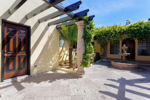 A courtyard with a wooden door, a column with climbing plants, a fountain, and a building with a blue dome in the background. - Villa Agave Azul | 5BR Ocean View Villa | Pool, Jacuzzi & Golf Views (Cabo del Sol)