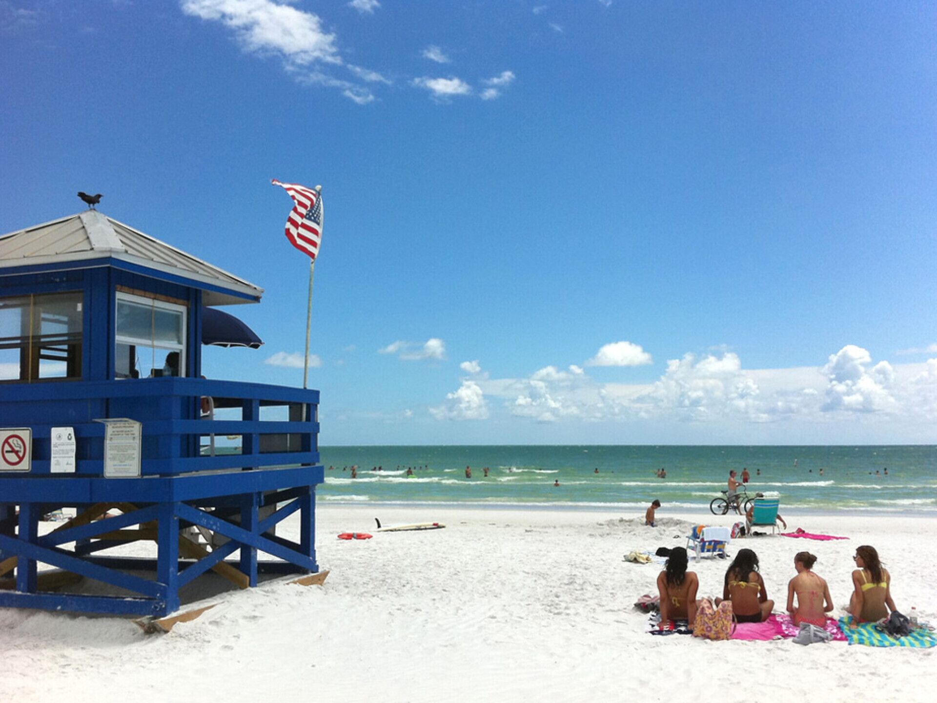 A beach lifeguard tower with an American flag, a sandy beach, and people relaxing on towels.