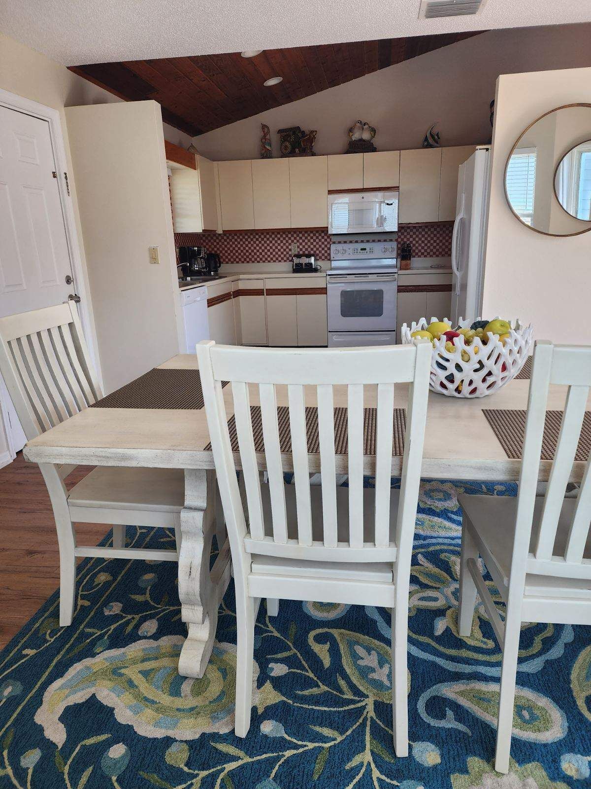 A compact kitchen with white cabinets, a microwave, and an oven. There is a dining table with two chairs and a fruit bowl.