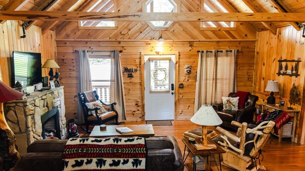 Ein gemütliches Blockhaushaus-Interieur mit Holzwänden, einem Steinkamin, einer bequemen Couch, einem Schaukelstuhl und einem Fernseher.