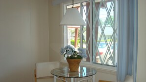 A small dining area with a glass table and two chairs, a wicker basket with flowers, and a window with a view of a street and parked cars. - Sea Drift Motel (Old Orchard Beach)
