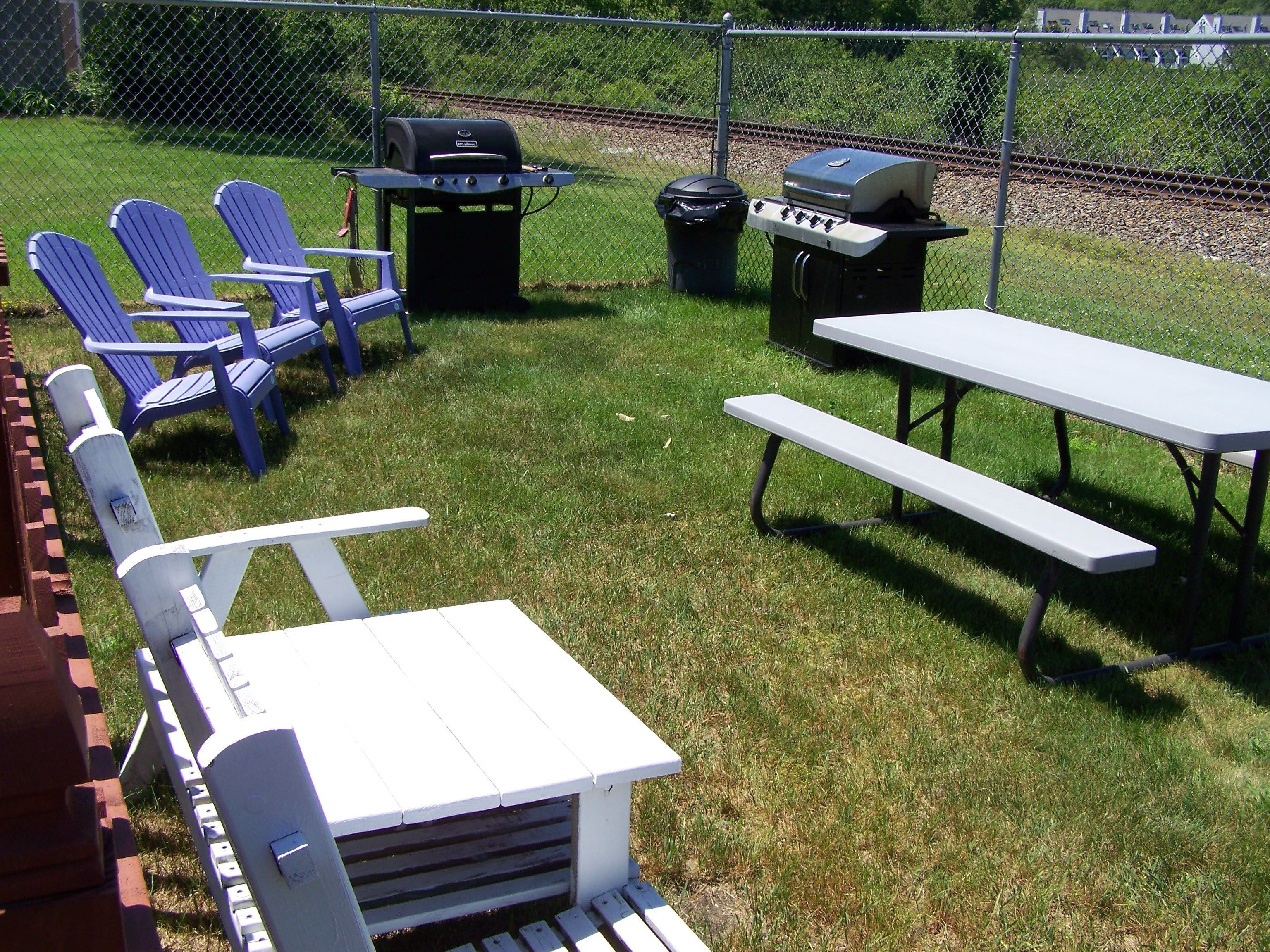 A backyard with a picnic table, blue Adirondack chairs, a grill, and a trash can.