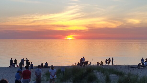 Eine Strandansicht, auf der Menschen am Sand sitzen und stehen und den Sonnenuntergang über dem Wasser betrachten.