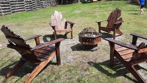 Ein hölzerner Feuerkorb, umgeben von vier Adirondack-Stühlen in einer grünen Außenanlage.