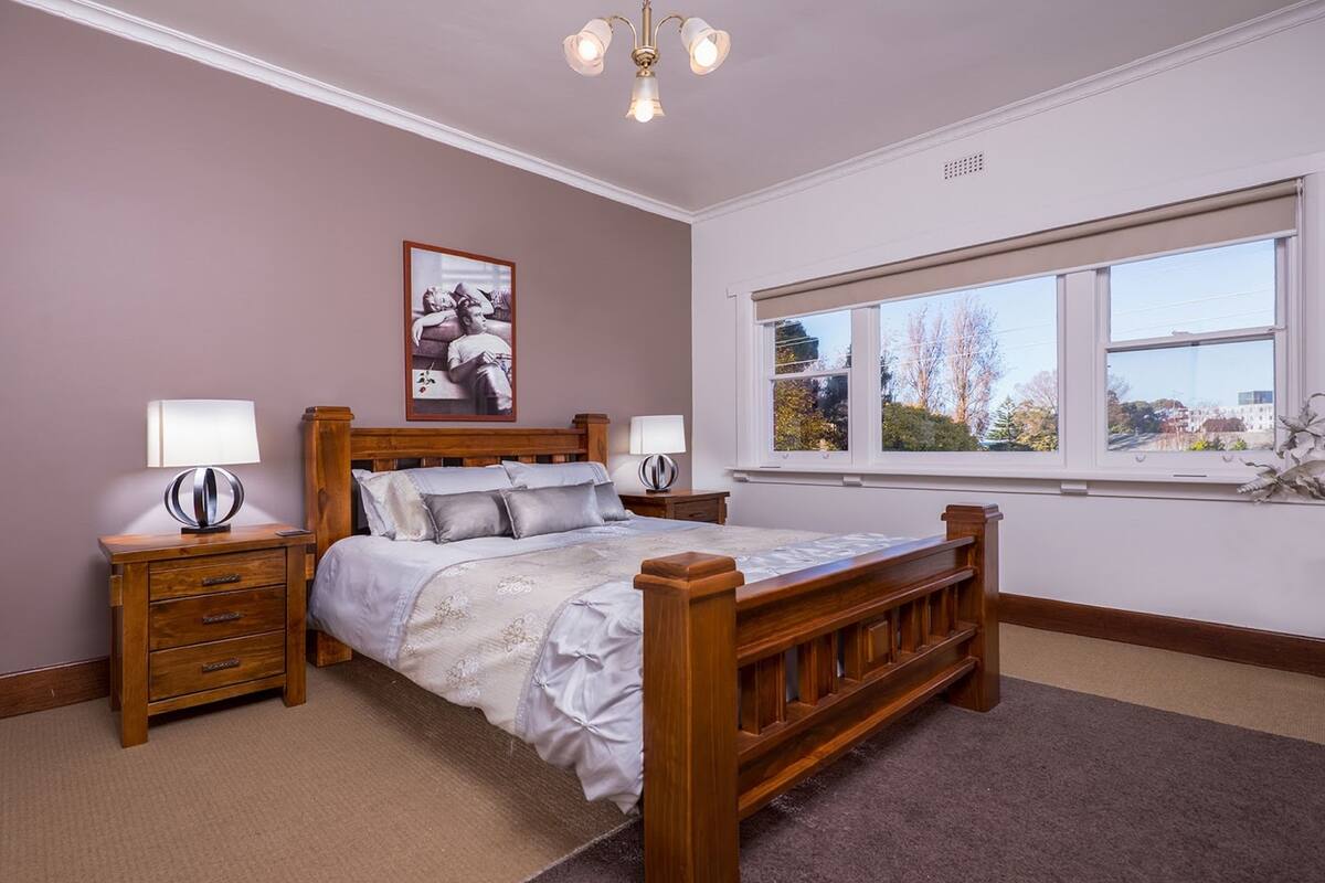 Een slaapkamer met een houten bed, twee nachtkastjes, een lamp en een groot raam met uitzicht, plus een plafondlamp.