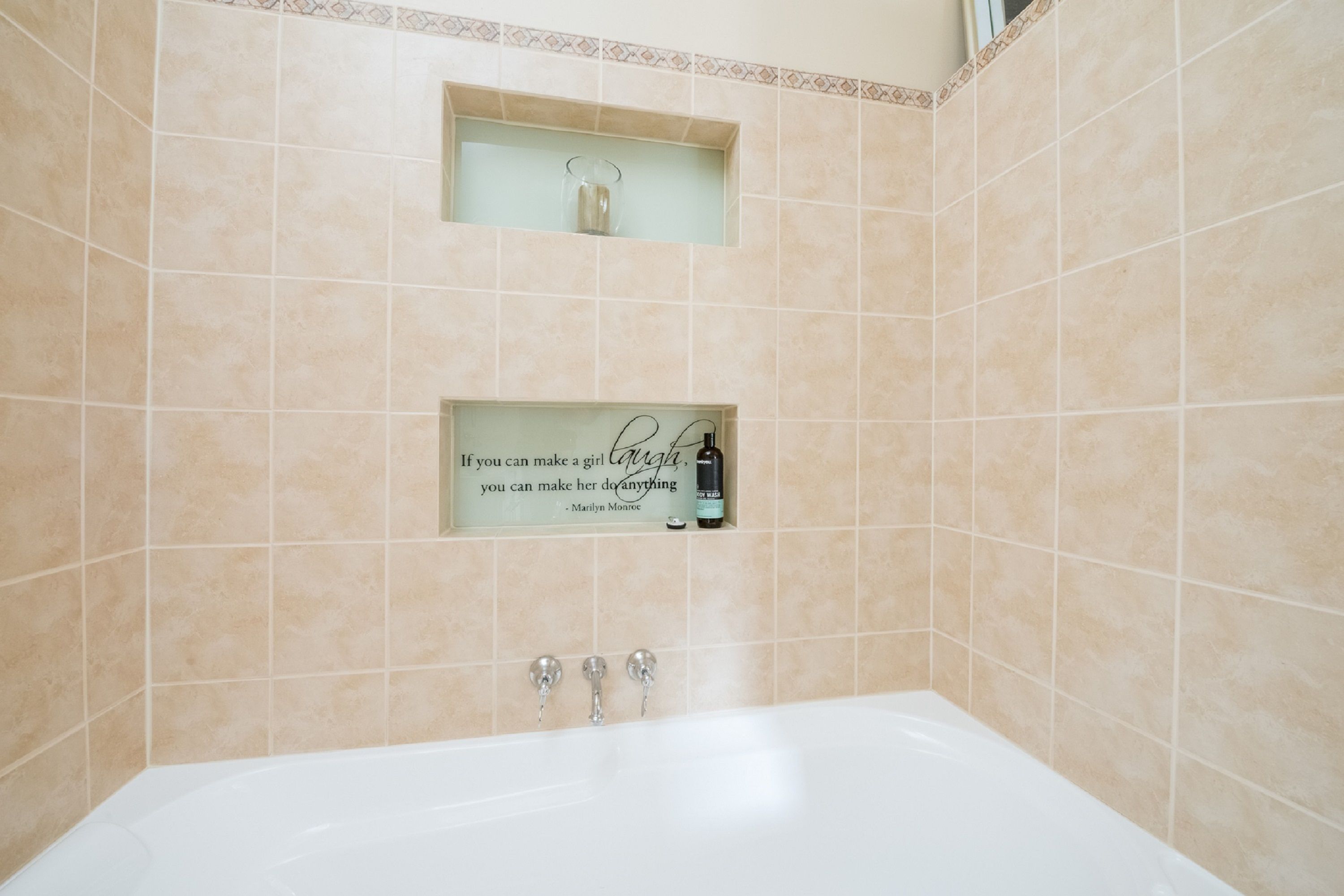 A bathroom with beige tiles, a white bathtub, and a quote sign on the wall.