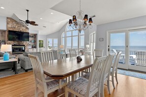 A spacious dining area with a large table and chairs, a chandelier, and a view of the ocean through large windows. - La Dolce Vita: Soundfront, Shared Pool, Hot Tub (Corolla)