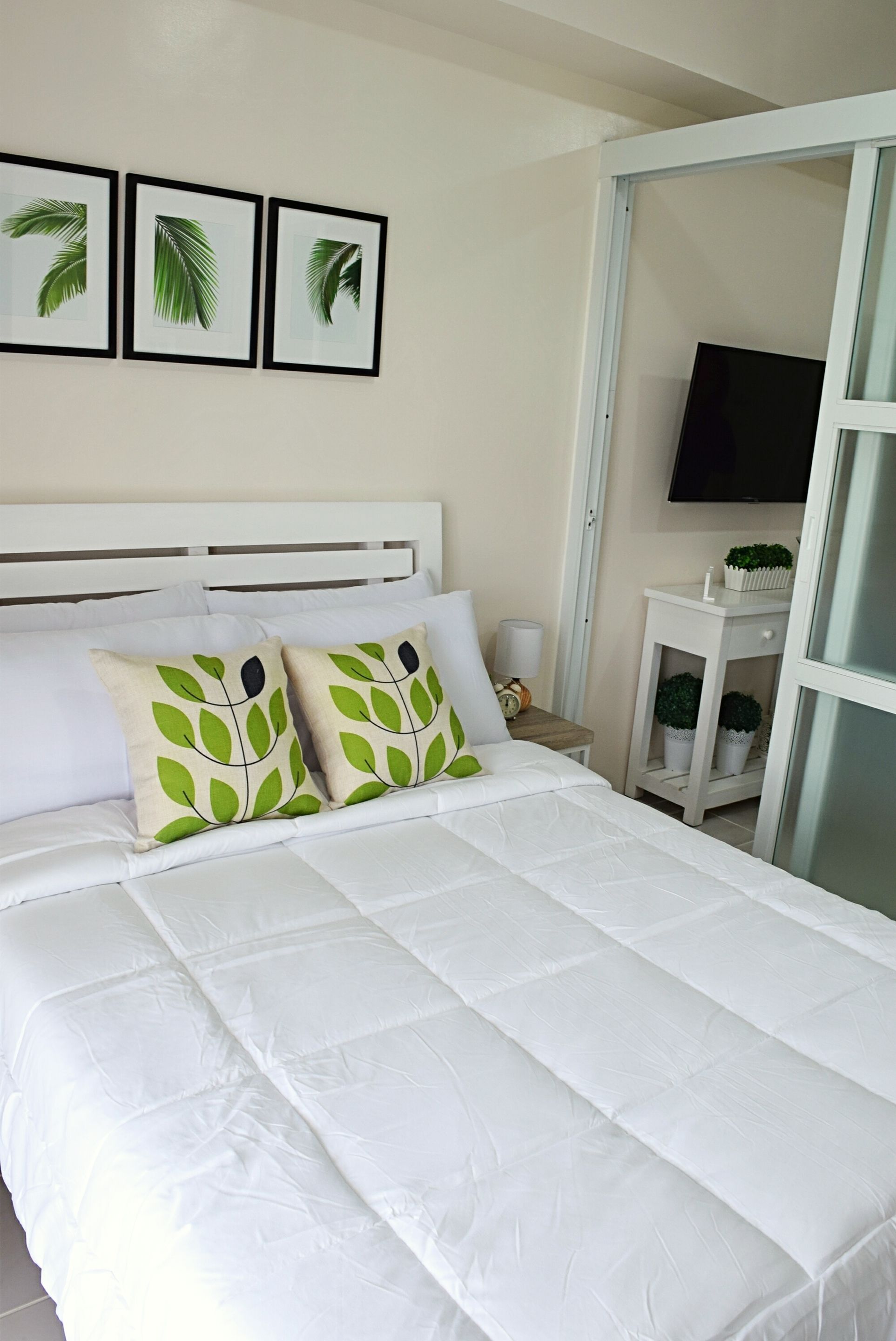 Una cama bien hecha con ropa blanca y almohadas con estampado de hojas verdes, una mesita de noche con una lámpara, y un televisor montado en la pared.