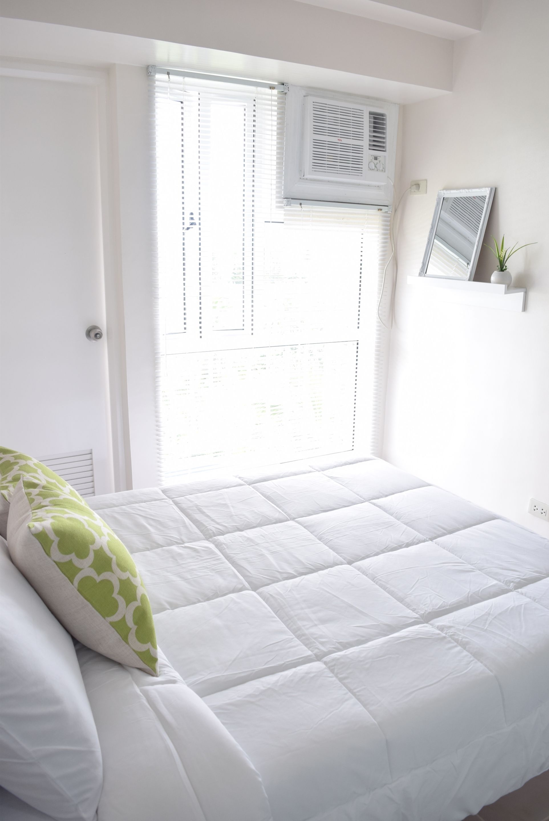 A bedroom with a bed, a window with blinds, an air conditioning unit, and a small shelf with a plant.