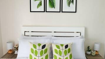 Una cama bien hecha con ropa blanca y dos almohadillas decorativas con estampado de hojas. Encima de la cama, hay tres láminas con motivos de hojas enmarcadas en la pared.
