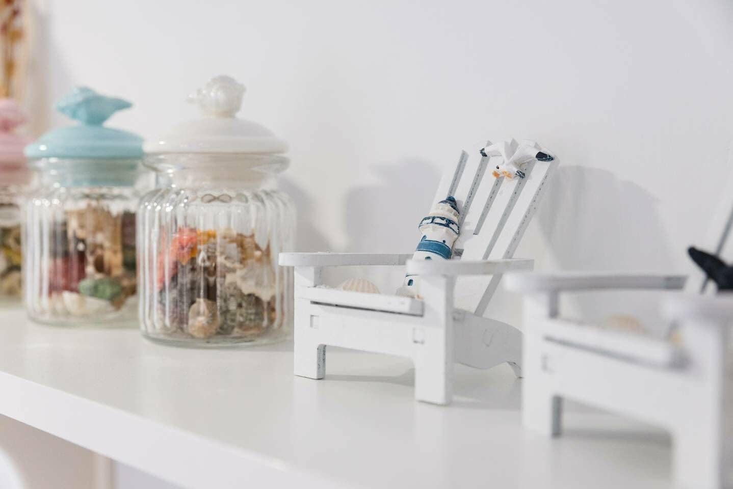 A white shelf with two glass jars containing various items and a small white chair with a decorative item on it.