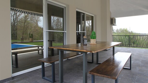 A patio with a wooden table and benches, large sliding glass doors, and a view of a pool and trees.