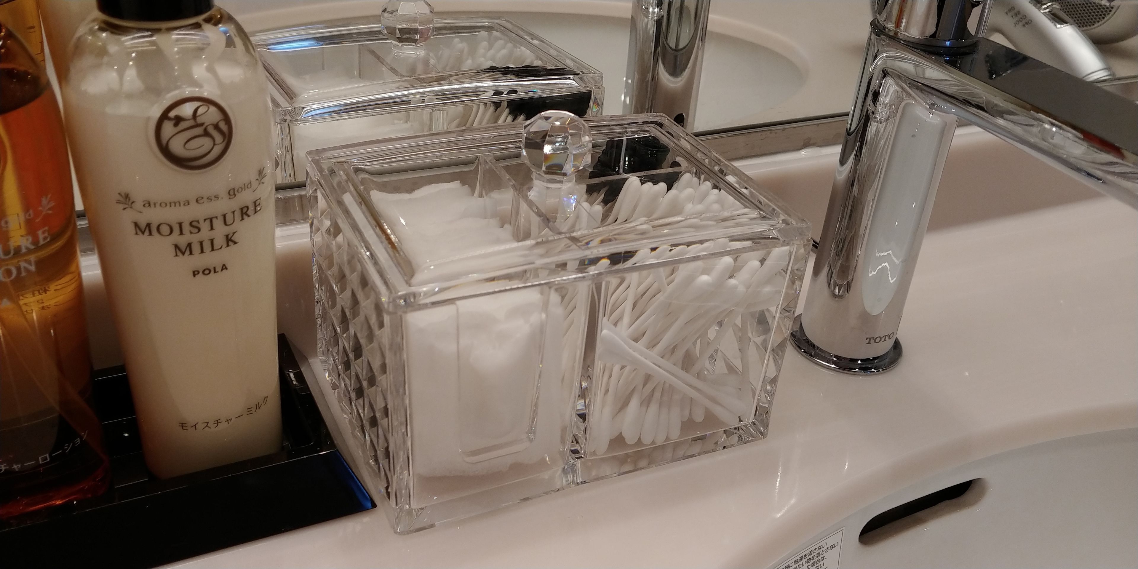 A bathroom sink with a bottle of Moisture Milk, a glass container with cotton swabs, and a Toto faucet.