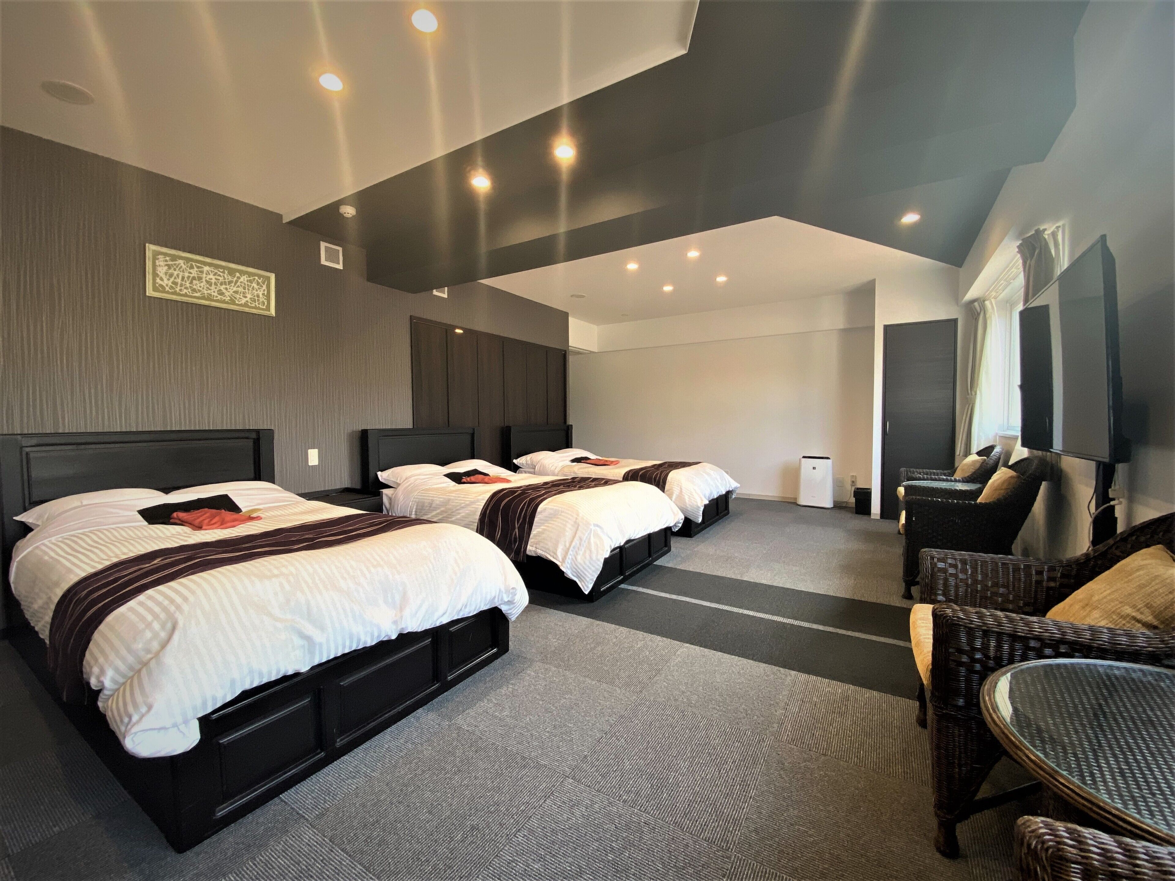A hotel room with three beds, a TV, a seating area, and a ceiling with recessed lighting.