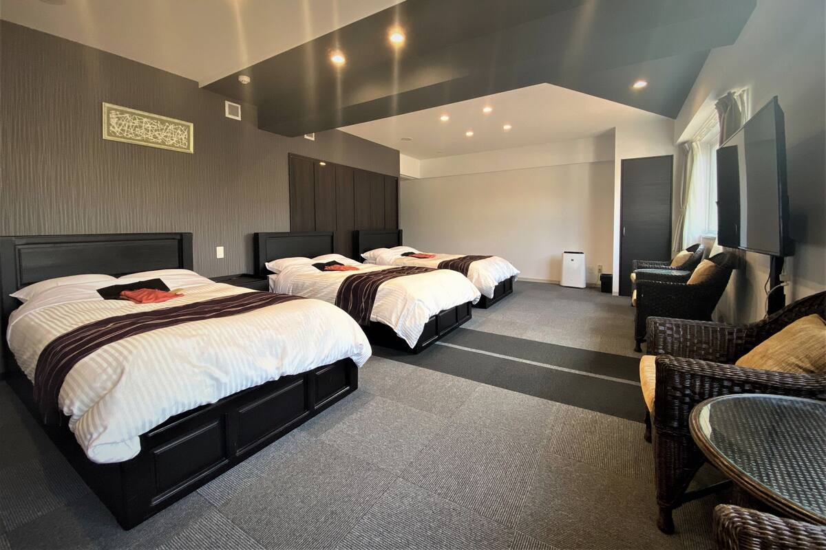 Habitación de hotel con tres camas, televisión, zona de estar e iluminación empotrada en el techo.