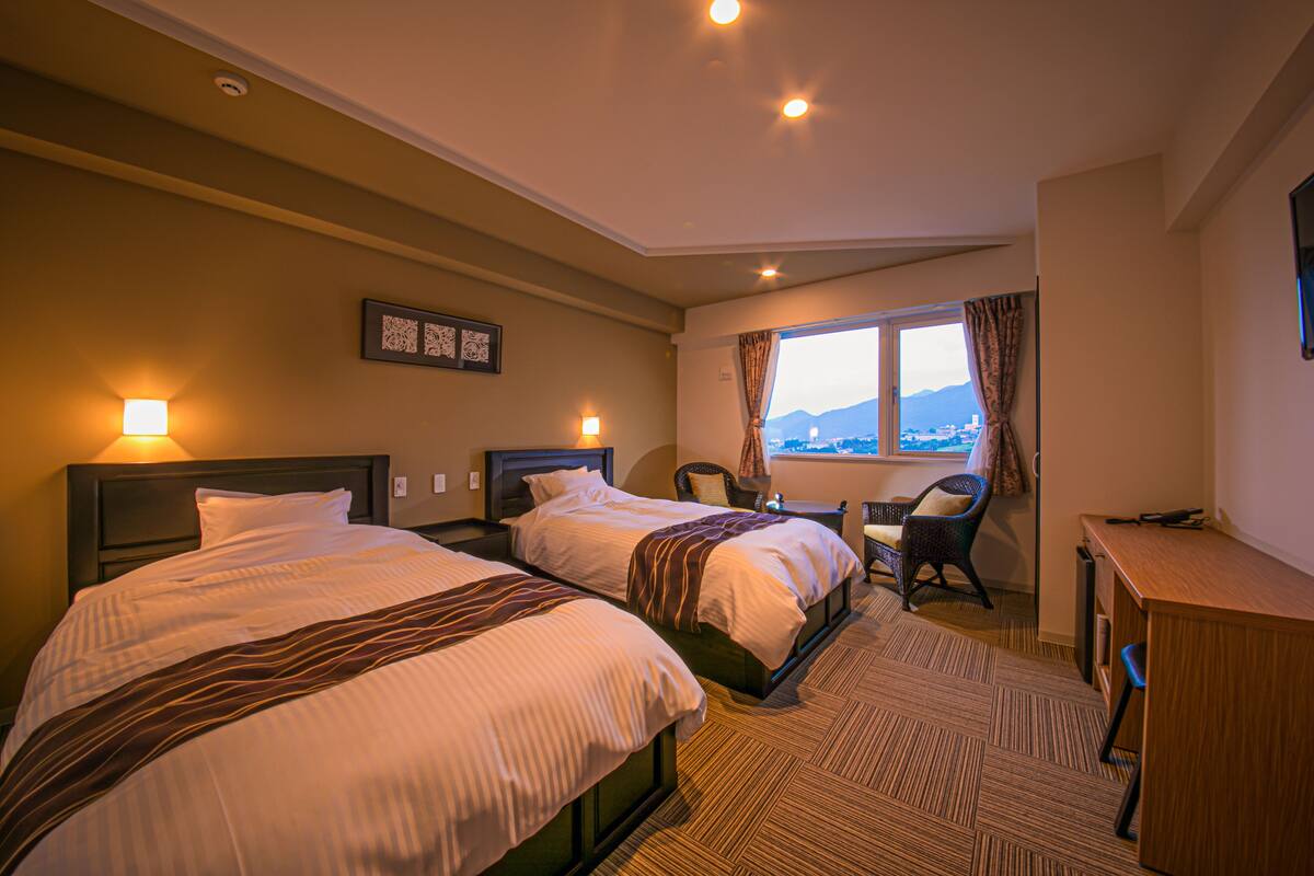 A hotel room with two beds, a desk, a chair, and a view of the city through the window.