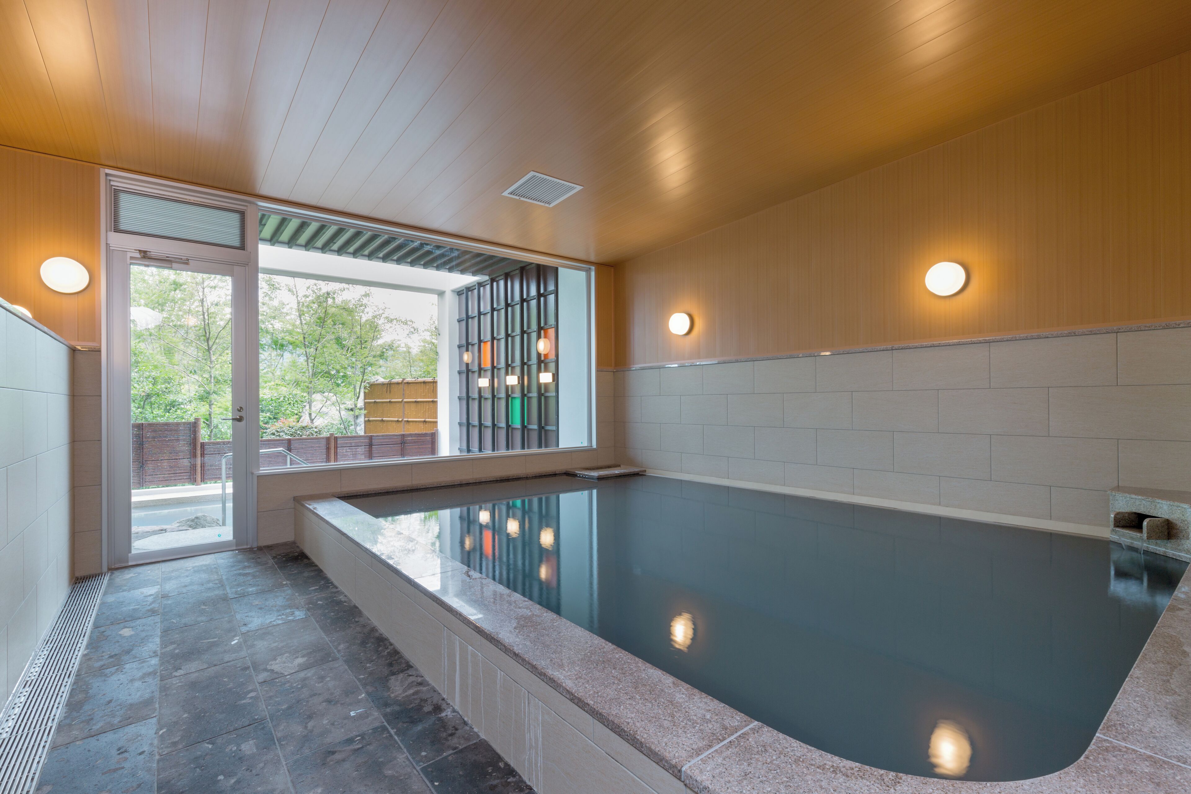 A modern indoor swimming pool with a clear view of the outside, a tiled floor, and a wooden ceiling.