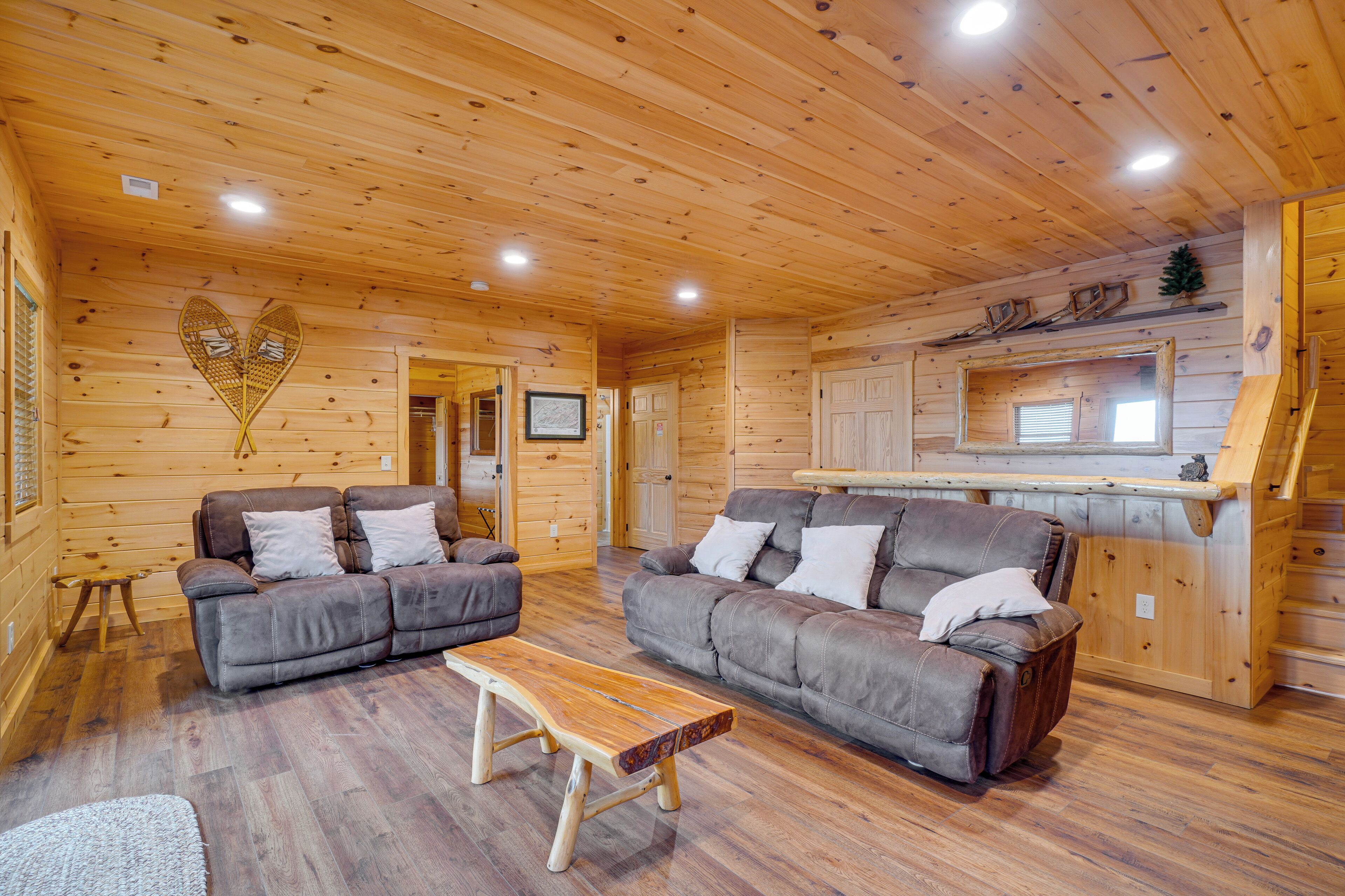 Interior de una cabaña de madera con dos sofás, una mesa de centro de madera y una zona de cocina con ventanal.