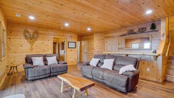 A wooden cabin interior with two sofas, a wooden coffee table, and a kitchen area with a window.