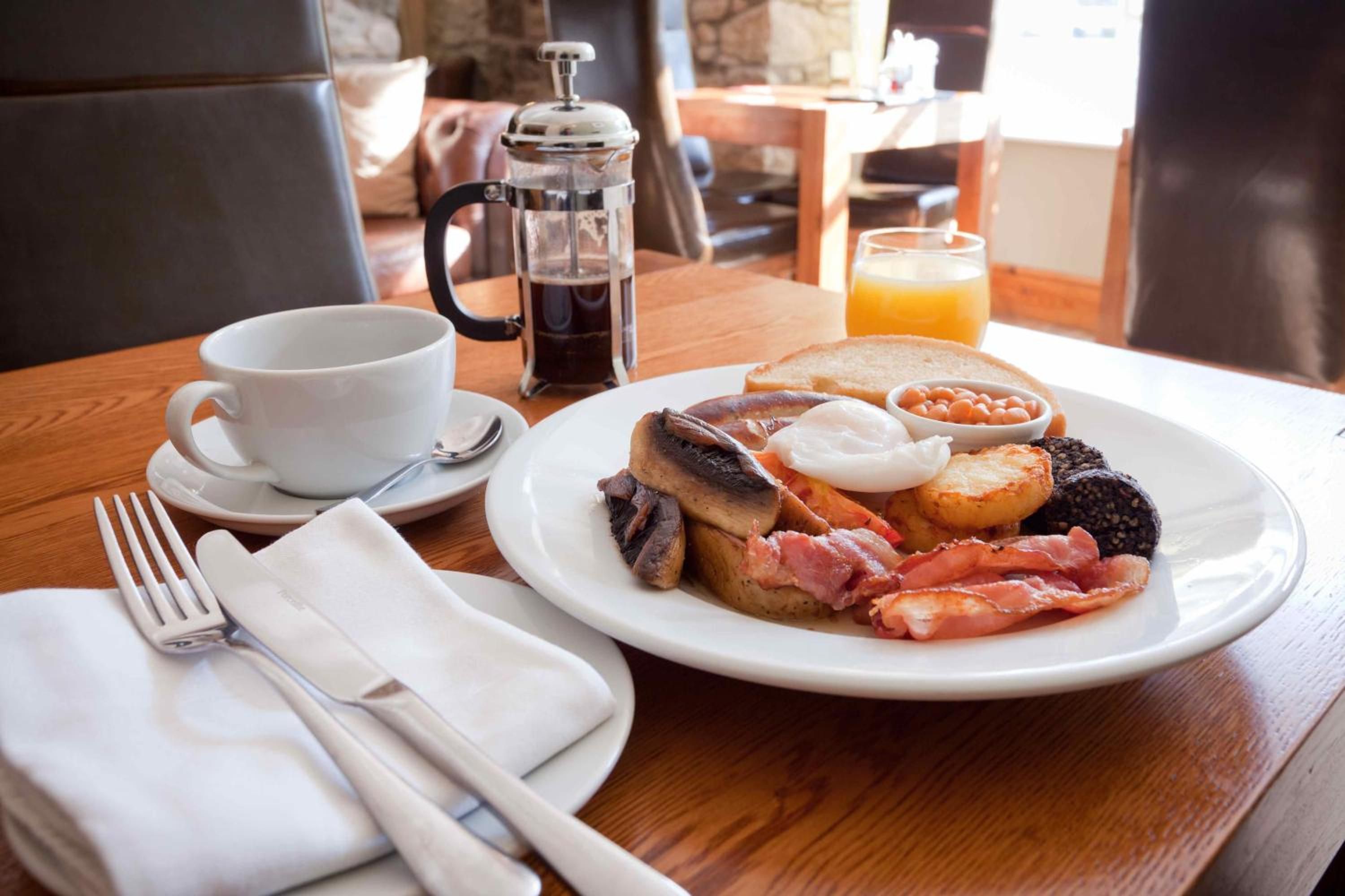 Une table en bois dressée pour le petit-déjeuner, avec une assiette contenant des œufs, du bacon, des champignons et des toasts, une cafetière, une tasse de café et un verre de jus d’orange.