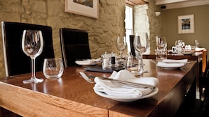 A dining table set with wine glasses, plates, and cutlery, with a stone wall and framed artwork in the background.