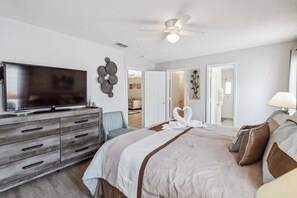 A bedroom with a bed, a TV, a dresser, a ceiling fan, and a door leading to another room.