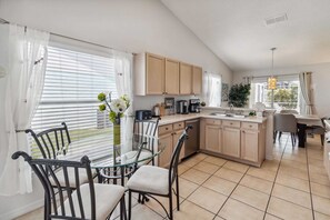 A modern kitchen with a dining area, featuring a glass table, metal chairs, and light wooden cabinets.