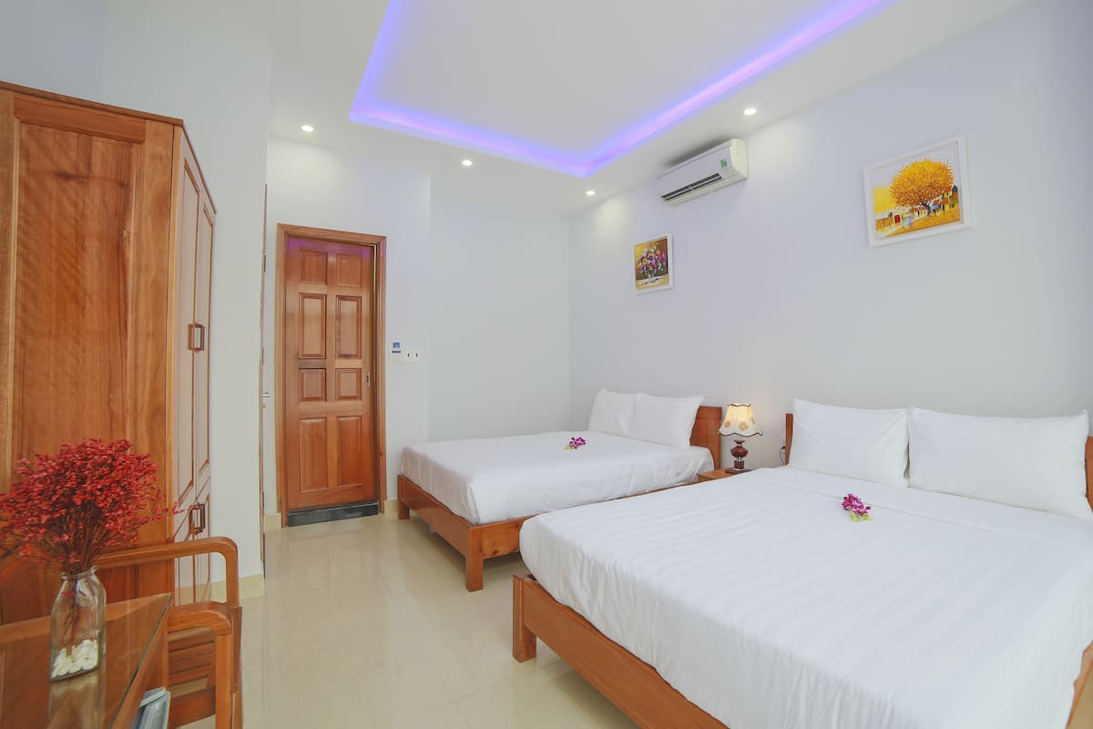 A hotel room with two beds, a wooden wardrobe, a dining table with chairs, and a vase of flowers.