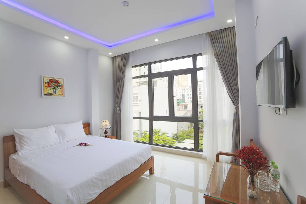 A hotel room with a bed, a TV, a window with curtains, a painting, and a vase with flowers.
