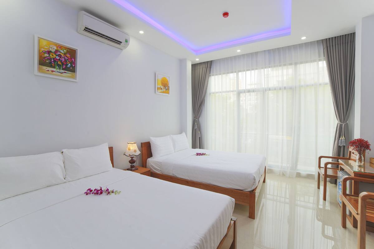 A hotel room with two beds, a desk, and a chair. There is an air conditioning unit on the wall and a window with curtains.