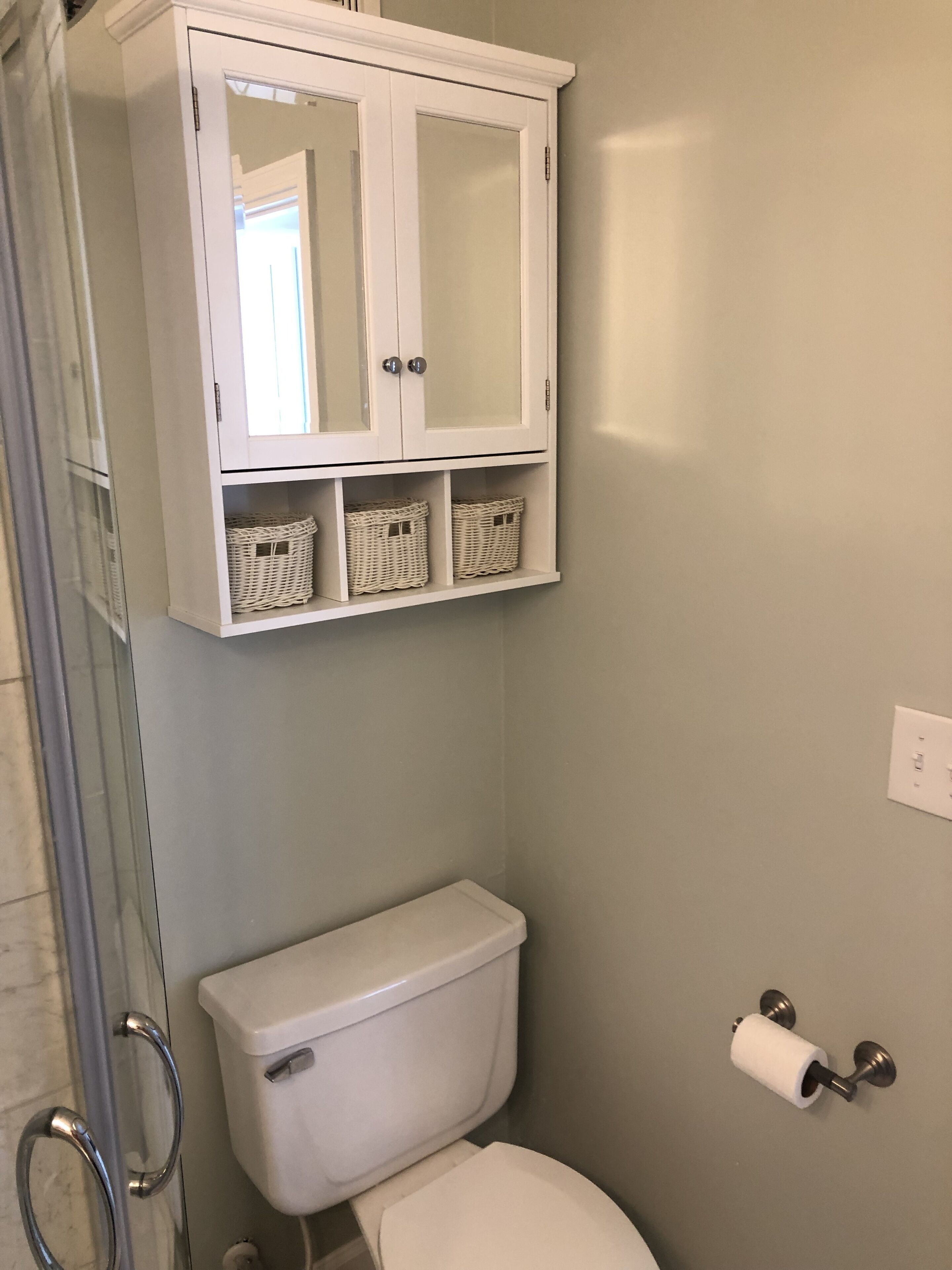A bathroom with a white toilet, a white cabinet with glass doors, and wicker baskets on the lower shelf.
