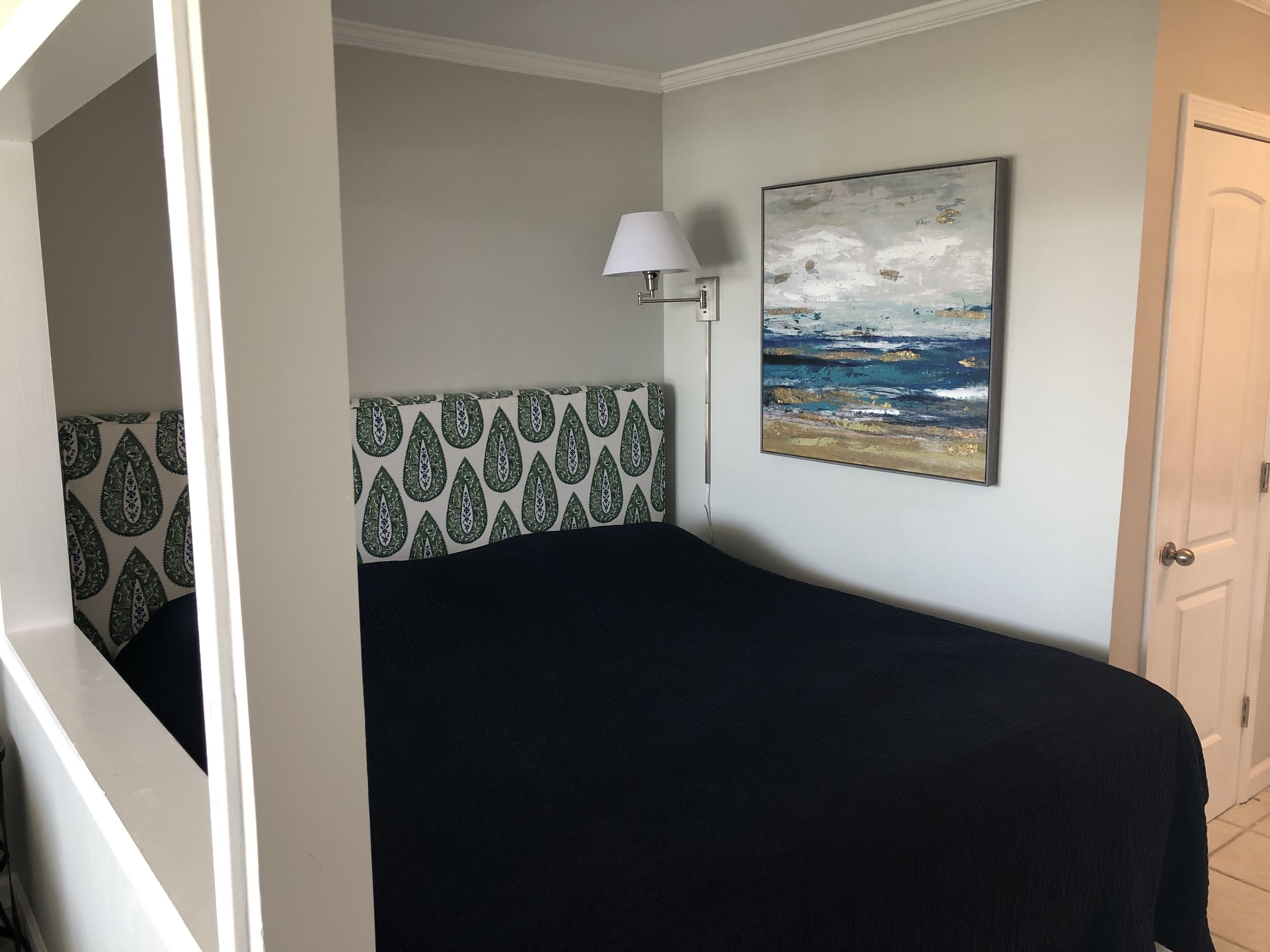 A bedroom with a bed, a patterned headboard, a painting of a beach scene, a bedside lamp, and a door.