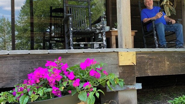 Une véranda en bois avec une chaise noire et un banc, un jardinier avec des fleurs roses et une pancarte sur laquelle on peut lire « RELAX ».
