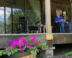 A wooden porch with a black chair and a bench, a planter with pink flowers, and a sign that reads "RELAX". - Cedar Cabin style house in Toledo Bend (Noble)
