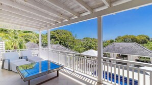 Un espacio exterior techado con una mesa de vidrio, muebles blancos y vistas a una piscina y vegetación.