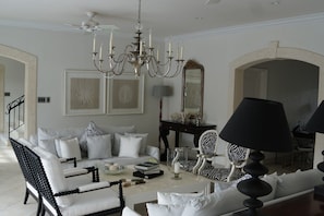 A living room with a white sofa, black and white armchairs, a chandelier, and a large mirror. - Tradewinds at Sandy Lane Estate | Timeless Luxury in Barbados’ Most Prestigious Address (Holetown)