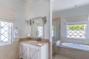 A bathroom with a large mirror, a sink, a bathtub, and a shower. There are windows with white curtains and a view of greenery outside.