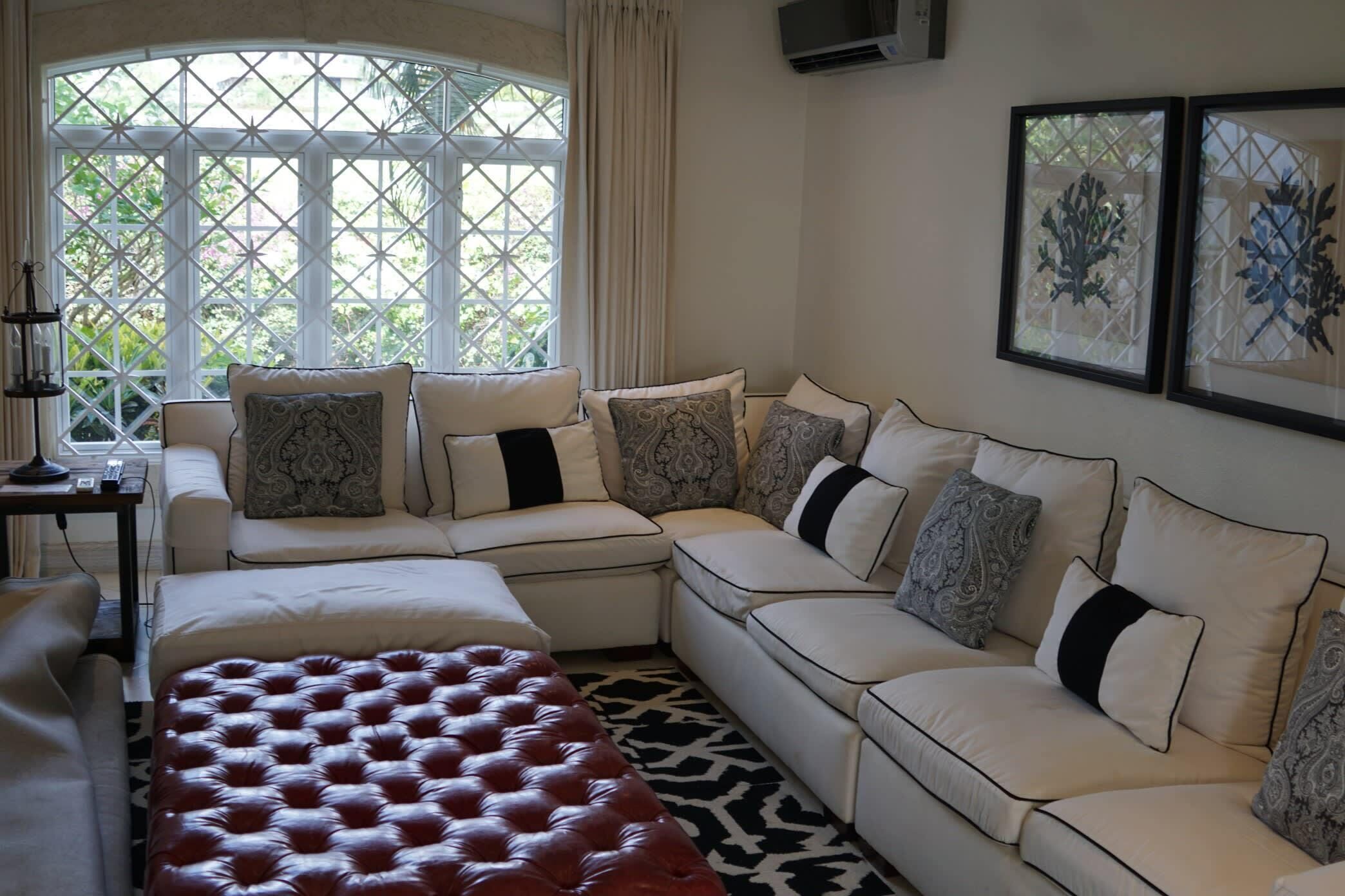 A modern living room with a sectional sofa, a red tufted ottoman, and a large window with a view of greenery.