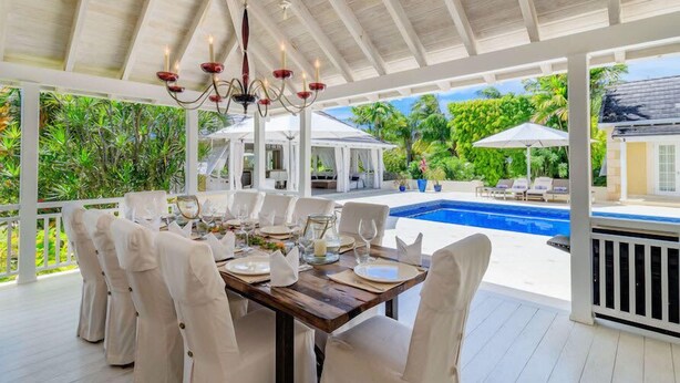Un área de comedor al aire libre con una mesa de madera dispuesta para seis personas, rodeada de sillas blancas, bajo un pabellón blanco con una piscina al fondo.
